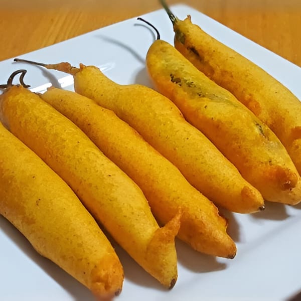 Six golden-fried chili peppers, known as Mirchi Bajji, arranged diagonally on a white plate.